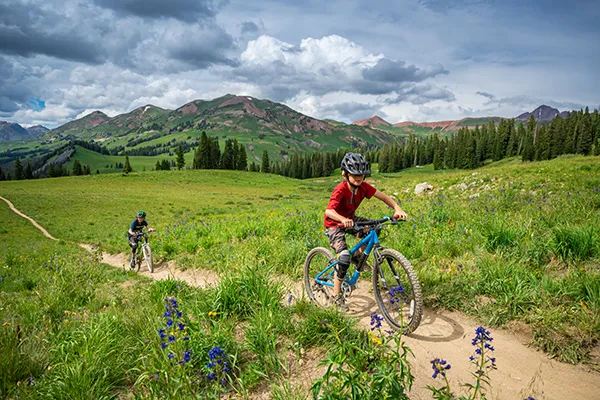 Two kids ride mountain bikes up a hill with mountains in the background
